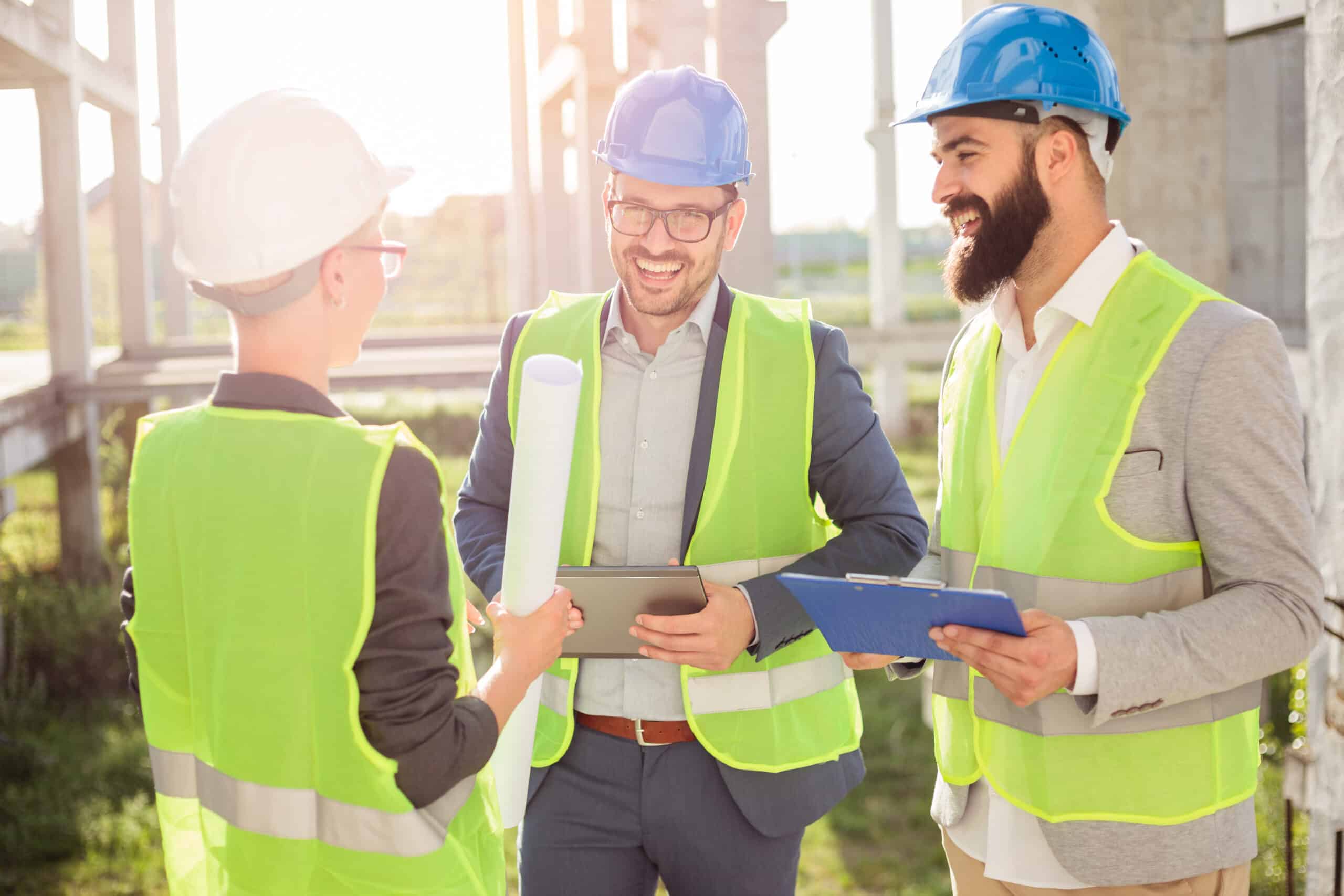 Three construction professionals in hard hats and safety vests discussing plans at a construction site, one holding a tablet and another with a clipboard.