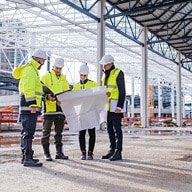 Five construction workers in hard hats and safety vests examining a large white blueprint at a construction site.